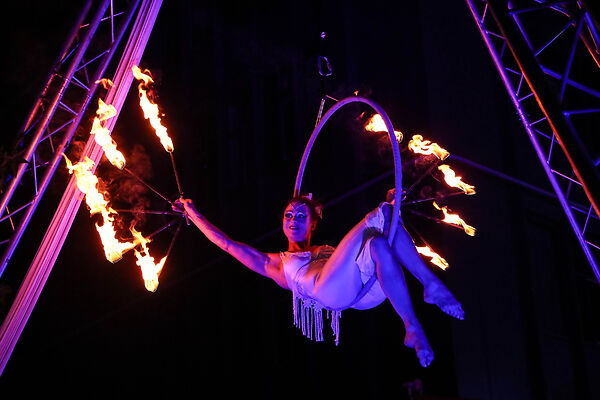 Auf dem Bild ist eine Akrobatin bei einer Feuershow zu sehen. Die Akrobatin sitzt auf einem in der freien Luft hängenden Reifen.
