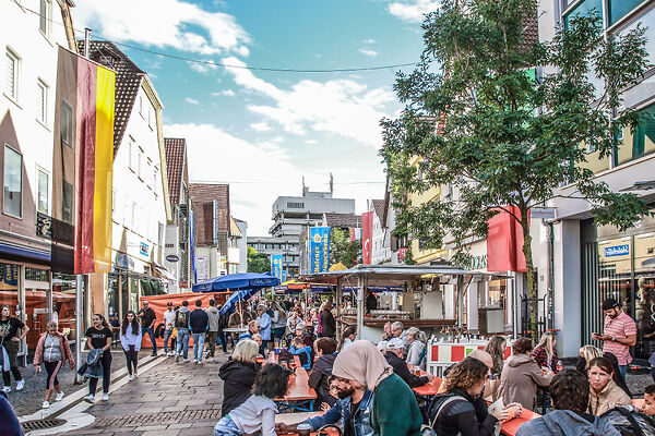 Auf dem Bild sind Menschen auf einem Stadtfest beim Essen und Unterhalten zu sehen. 