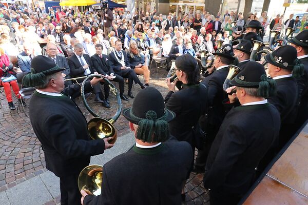 Auf dem Bild sind Musikanten zu sehen, die auf Hörnern musizieren. Eine Menschenmenge sitzt vor ihnen und hört ihnen zu.