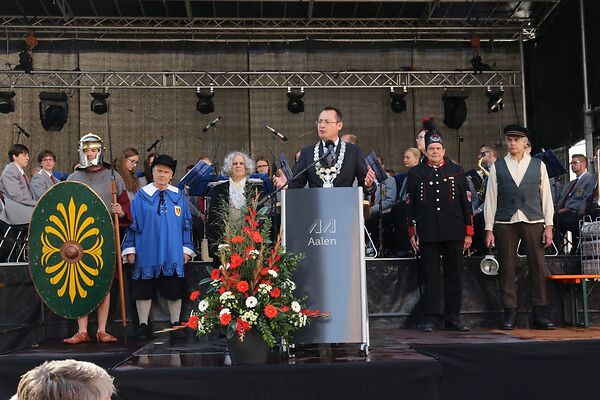 Auf dem Bild ist der damalige Oberbürgermeister Thilo Rentschler auf einer Bühne zu sehen. Hinter ihm stehen in verschiedenen Gewändern gekleidete Personen.