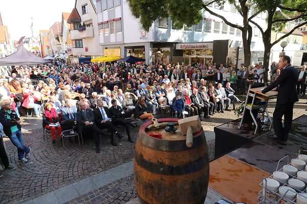 Im Vordergrund des Bildes ist ein Bierfass zu sehen. Im Hintergrund ein Publikum, das der Rede vom damaligen Oberbürgermeister Thilo Rentschler zuhört.