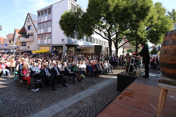 Auf dem Bild ist ein auf Stühlen sitzendes Publikum zu sehen, das einem Sprecher am Rednerpult zuhört.