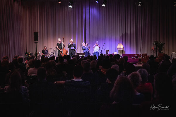 Auf dem Bild sind ein Publikum von hinten und fünf Musiker auf der Bühne zu sehen.