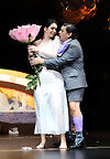 Auf dem Bild sind zwei Opernsänger*innen zu sehen. Er umarmt sie, sie hält ein übergroße Blume in der Hand.