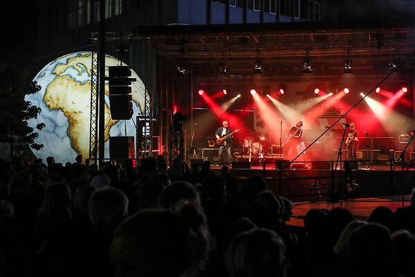 Auf dem Bild ist eine Band zu sehen, die auf einer in rot beleuchteten Bühne vor großem Publikum auftritt.