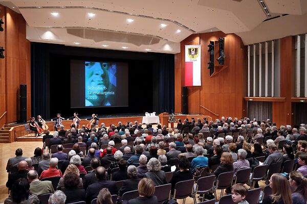 Auf dem Bild ist das Publikum des Festaktes in einer FEsthalle zu sehen. Auf der Bühne tritt ein Cello-Orchester auf.