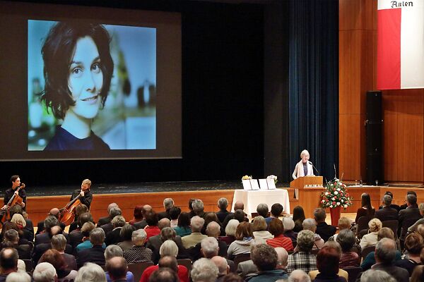 Auf dem Bild ist eine Dame an einem Rednerpult zu sehen, die zu ihrem Publikum spricht. Im Hintergrund ist das Foto einer Siegerin des Schubart-Literaturpreises auf einer Leinwand zu sehen.