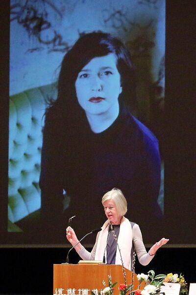Auf dem Bild ist eine Dame an einem Rednerpult zu sehen, die zu ihrem Publikum spricht. Im Hintergrund ist das Foto einer Siegerin des Schubart-Literaturpreises auf einer Leinwand zu sehen.