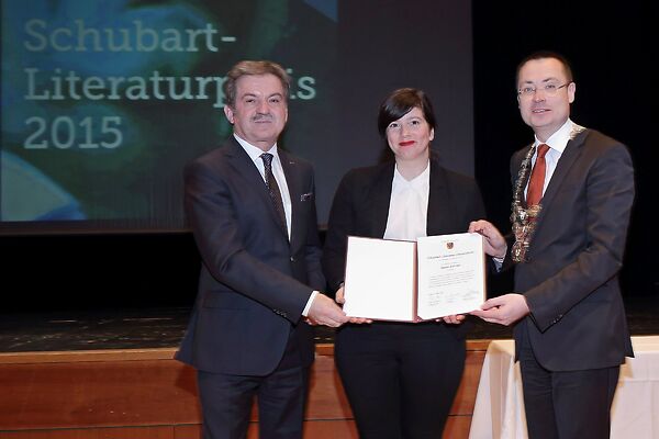 Auf dem Bild ist eine der Schubart-Literaturpreis-Trägerinnen zu sehen, die mit zwei HErren eine Ukrunde in die Kamera zeigt.