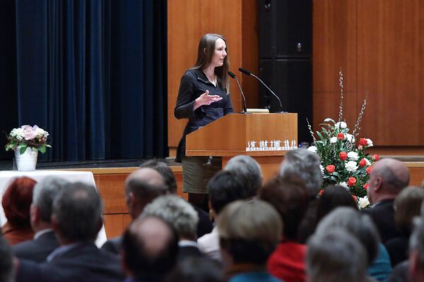 Auf dem Bild ist eine Frau hinter einem Rednerpult zu sehen, die zu ihrem Publikum spricht.