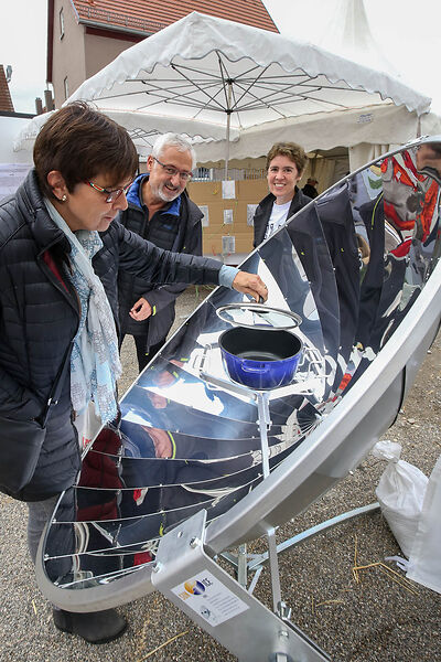 Auf dem Bild sind zwei Frauen und ein Mann zu sehen. Vor ihnen steht eine Art Satellitenschüssel, in der ein Topf steht. EIne der Frauen hebt den Deckel des Topfes.