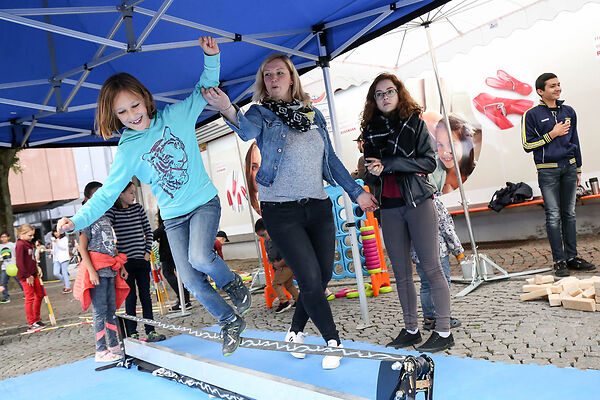 Auf dem Bild ist ein auf einer Slackline tanzendendes Kind zu sehen. Zwei Frauen stehen daneben und eine gibt dem Kind Hilfestellung.