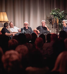 Auf dem Bild sind fünf Männer der Band Stumpfes Zupf & Ziehkapelle auf der Bühne sitzend auf einem Sofa zu sehen. Im Hintergrund sind Musikinstrumente zu sehen.