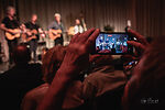 Auf dem Bild sind 5 Männer auf der Bühne vor Publikum auf einem Smartphonebildschirm zu sehen.